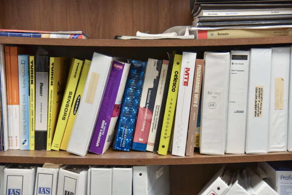 Industrial automation manuals, sensor guides, and technical reference binders organized on office shelves.